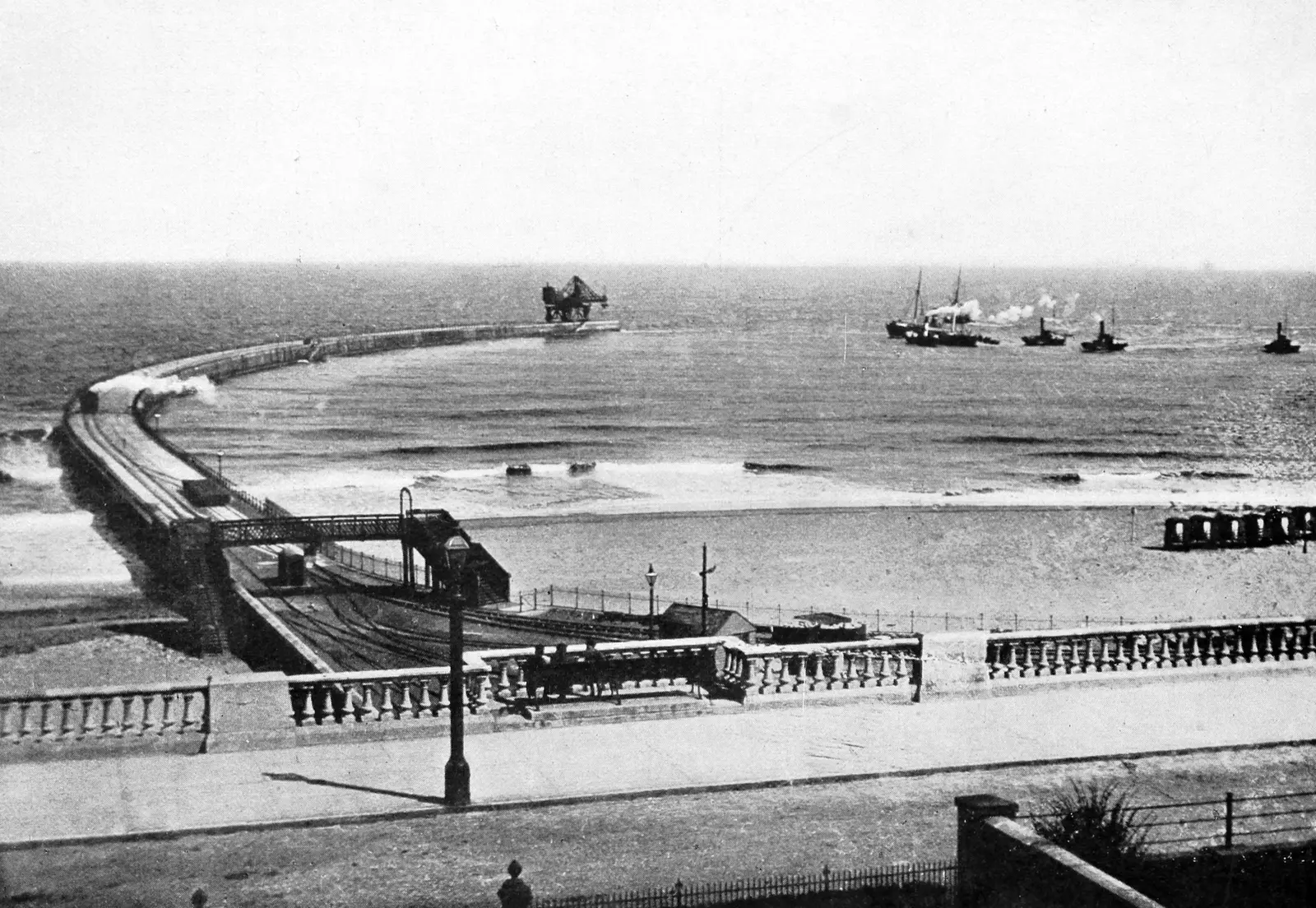 Roker Pier curving out to sea beyond the balustraded promenade, with a steam train on the pier, a crane at its end and tugs on the water.